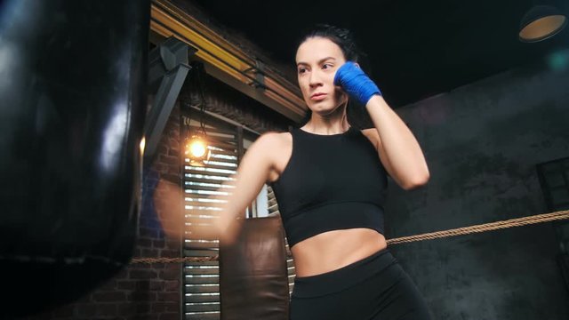 Attractive fit boxer woman with perfect slim body hitting punching bag at dark gym interior