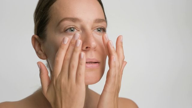 Close-up beauty portrait of young woman with smooth healthy skin, she gently touches her face with her fingers on white background and smiles