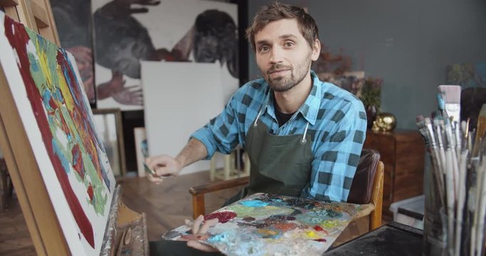 Young handsome male artist painting picture in cozy studio using palette knife technique, abstract style