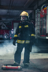 Fototapeta premium Portrait of a female firefighter wearing a helmet and all safety equipment a while holding a tomahawk and wearing an oxygen mask indoors surrounded by smoke