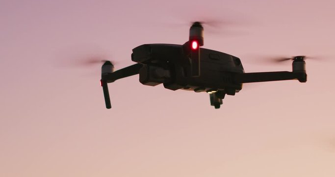 Silhouette of a quadcopter drone hovering and turning in a pink sky at dusk with blinking landing signal lights