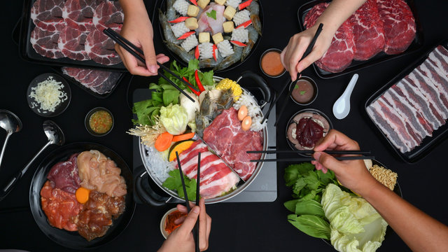 Top view of people eating Shabu-Shabu in hot pot with fresh sliced meat, sea food, and vegetables with black background