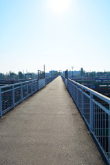 A narrow gray reinforced concrete bridge over a junction of railways and freight trains lit by the bright sun with blue sky.