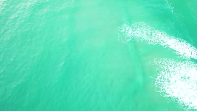 Drone Above Turquoise Blue Ocean Waves Crashing Onto The Shoreline With White Wash On A Sunny Day