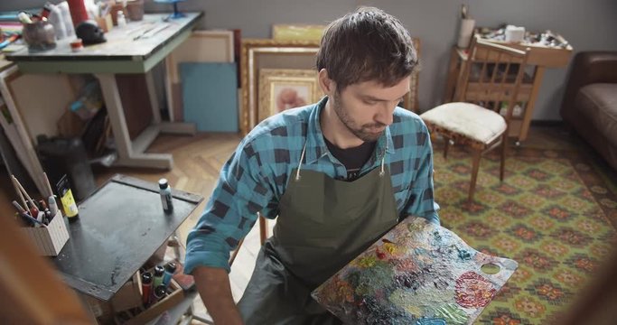 Young bearded man in apron and casual blue shirt painting picture in artist studio, hobby
