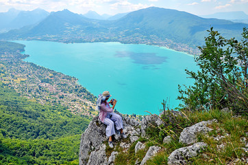 A young woman sitting on the rock and admirring a panoramic viewview on the Lake Annecy from mont Veyrier to mont Baron hiking track, France