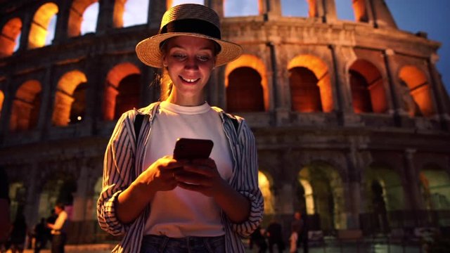 Attractive female tourist in hat standing in Rome near historical place laughing at funny content from social network on mobile phone. Hipster girl text messaging via application on smartphone
