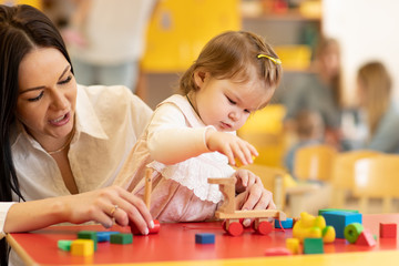 Little caucasian child girl playing with Montessori toy in pre-school. Woman teaching kid in kindergarten