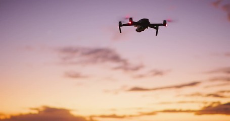 Orbiting view of a quadcopter drone silhouette hovering and flying at sunset with a purple yellow sky - Powered by Adobe