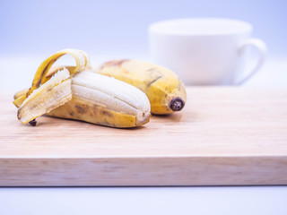 Closeup of couple yellow cultivated banana on wooden cutting board, blurry coffee cup and gray background in the kitchen for healthy eating or dieting concepts and ideas.