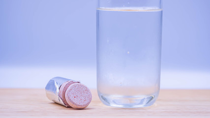 Closeup of multivitamin effervescent tablet pills with a glass of cold water on wooden table and gray blurry bedroom background.