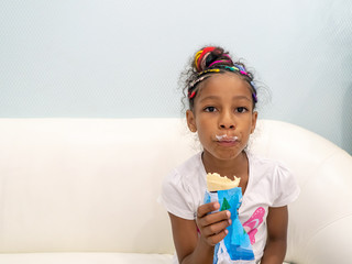 Jamaican European ethnicity little girl with colored braids sitting on a white sofa inside the house eating ice cream