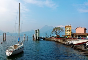 Fototapeta na wymiar Lakeside scenery of Varenna in springtime, a beautiful village by Lake Como in Lombardy, Italy with a view of sailboats parking at the pier by lakeshore & snow capped mountains in distant background
