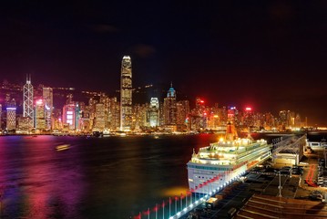 Obraz premium Night scenery of Hong Kong with a skyline of crowded skyscrapers by Victoria Harbor, a luxury cruise liner parking by the pier of Harbour City and lights of high-rise buildings glistening in twilight