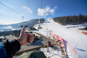 Human hand with thumb-up gesture on background of mountain ski-lift rope way on sunny winter day. Moving cable cars, bright blue sky and dark spruce trees, view from above. Active lifestyle concept.