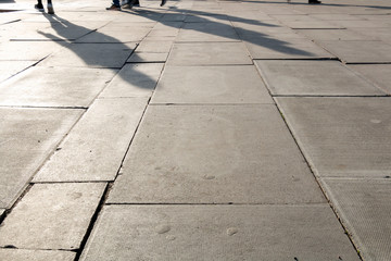 Black and white Picture of footpath as many people walk cross by. People on the street. Picture focusing on concrete with pedestrian’s shadow on. Silhouettes and Shadows of a person on the street.