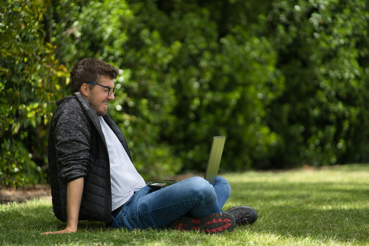 Young man looking at his computer in a park. He is sitting on the green grass and seems having fun. Natural Environment
