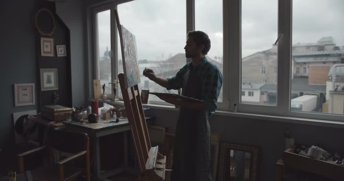 Young male painter holding palette painting picture in front of window, human silhouette