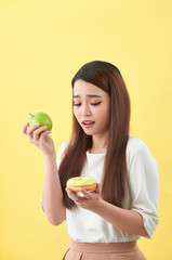 Portrait of a smiling young asian woman choosing between donut and green apple isolated over yellow background