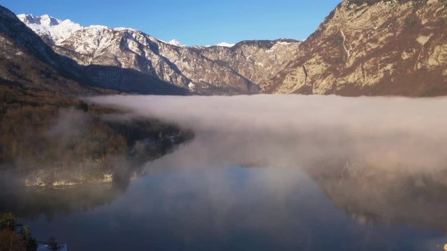 Flying over low fog covering lake in Slovenia viewing the mountains on sunny day.
