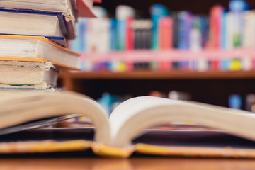 Textbook stack on wooden table with blur background of bookshelf and opened book in library. Photo concept of education, knowledge, traditional learning, and literature. Selective Focus