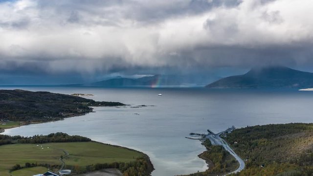 A ferry crossing from Bognes to Lodingen. The fjord is calm, thick white clouds hanging above, a rainbow appears in a distance, HIgh mountains and green fields are framing the scene.