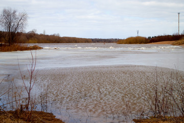 Spring landscape with river, yellow grass on the shore, trees without leaves and blue sky with white clouds in the background