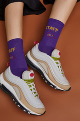 Cropped shot of a girl's foots in white sneakers, lying on a brown background. It is violet artwork socks with lettering "staff only" on foots. 