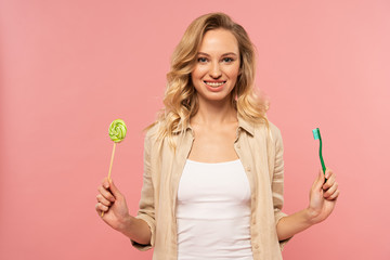 Smiling blonde woman holding toothbrush and lollipop isolated on pink