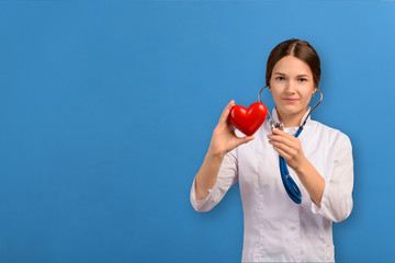 Doctor young girl in white coat with stethoscope examining red heart isolated on blue background. Concept for layout on topic of medicine and cardiology.