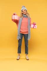 Smiling woman in hat and scarf holding gift boxes on yellow background