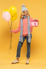 Smiling woman in hat and scarf holding balloons and looking at gift box on yellow background