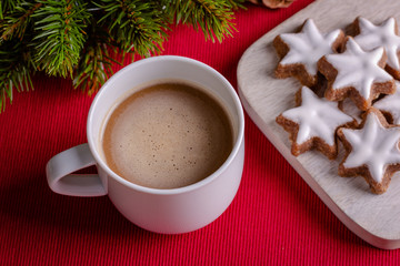Cup of coffee on red textile background with twig of decorative christmas tree and christmas cookies (cinnamon stars, Zimtsterne in German). Selective focus, top view, close-up. Christmas wallpaper.