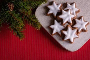 Christmas cookies (cinnamon stars, Zimtsterne in German) on wooden plate on red textile background with twig of decorative christmas tree. Selective focus, top view, close-up. Christmas wallpaper.
