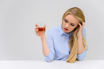 Concept woman with alcohol problems sits at a table with whiskey in a glass. Portrait of a beautiful blonde girl with excellent makeup with long smooth hair on a white background in a blue shirt.