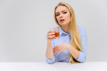 Concept woman with alcohol problems sits at a table with whiskey in a glass. Portrait of a beautiful blonde girl with excellent makeup with long smooth hair on a white background in a blue shirt.