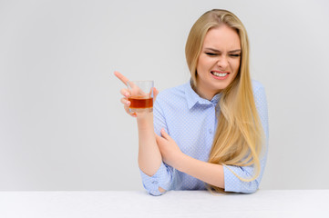 Concept woman with alcohol problems sits at a table with whiskey in a glass. Portrait of a beautiful blonde girl with excellent makeup with long smooth hair on a white background in a blue shirt.