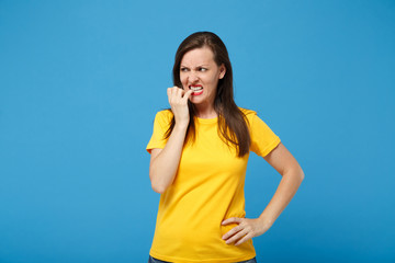 Nervous angry young brunette woman girl in yellow t-shirt posing isolated on bright blue wall background, studio portrait. People lifestyle concept. Mock up copy space. Gnawing nails, looking aside.
