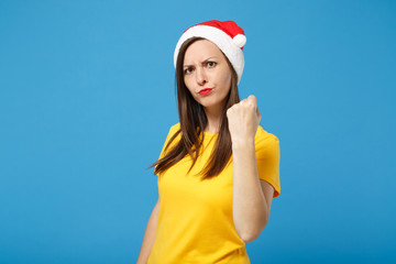 Angry strict young brunette woman Santa girl in yellow t-shirt Christmas hat posing isolated on bright blue background. New Year 2020 celebration holiday concept. Mock up copy space. Clenching fist.