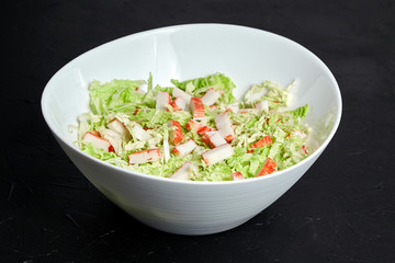 Chopped crab sticks and fresh chinese cabbage leaves in white ceramic bowl on black stone background. Vegetable and imitation crab meat, sea food.  Cooking salad