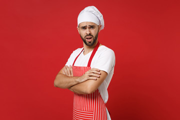Irritated young bearded male chef cook baker man in striped apron white t-shirt toque chefs hat posing isolated on red wall background. Cooking food concept. Mock up copy space. Holding hands crossed.