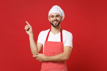 Smiling young bearded male chef cook baker man in striped apron white t-shirt toque chefs hat posing isolated on red background. Cooking food concept. Mock up copy space. Pointing index finger aside.