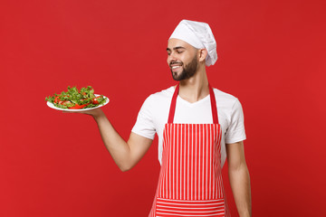 Smiling young bearded male chef cook or baker man in striped apron toque chefs hat posing isolated on red wall background. Cooking food concept. Mock up copy space. Holding plate with vegetable salad.