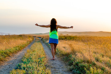 A young girl is walking along the road in a field in summer.Greeting to the sun.
