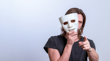 A long-haired man in a gray T-shirt looks out from behind a white mask and points his finger at the camera. The concept is you next, fraud. Copy space. Gender or split personality. Caution Warning.