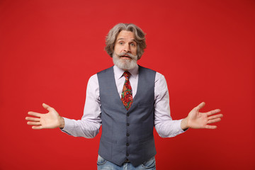 Naklejka premium Bewildered confused elderly gray-haired mustache bearded man in classic shirt vest colorful tie isolated on red background in studio. People lifestyle concept. Mock up copy space. Spreading hands.