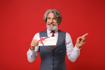 Smiling elderly gray-haired mustache bearded man in classic shirt vest and tie isolated on red background. People lifestyle concept. Mock up copy space. Holding gift certificate pointing finger aside.