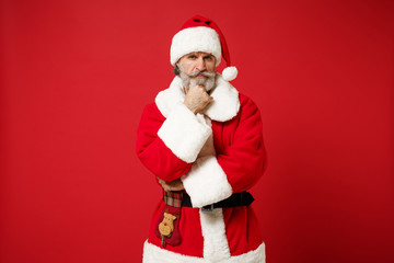 Serious elderly gray-haired mustache bearded Santa man in Christmas hat posing isolated on red background. New Year 2020 celebration holiday concept. Mock up copy space. Putting hand prop up on chin.