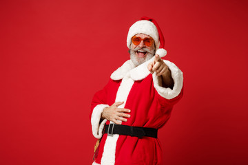 Fototapeta na wymiar Laughing elderly gray-haired mustache bearded Santa man in Christmas hat sunglasses posing isolated on red background. New Year 2020 celebration concept. Mock up copy space. Pointing finger on camera.