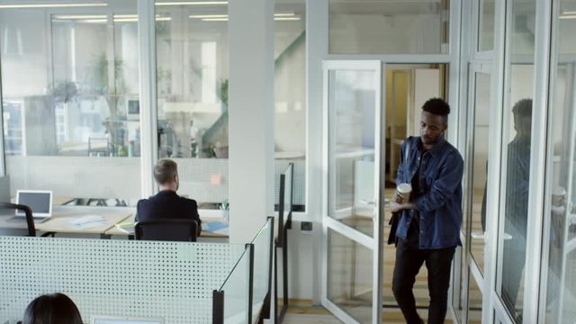 Tilt down of african american businessman walking with disposal coffee cup into modern open space office while colleagues working on laptops at desks